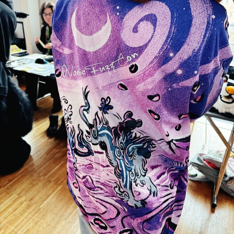 A person is wearing a colorful shirt with a mystical design. The shirt features a crescent moon and swirling patterns in shades of purple, pink, and blue. There is an artistic depiction of a creature resembling a dragon or lion, surrounded by floral designs. The text Nordic Fuzz Con is written across the back. In the background,