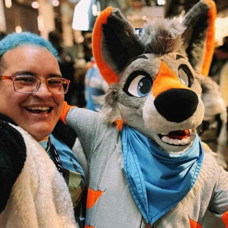 A smiling person with blue hair and glasses is taking a selfie, posing arm-in-arm with someone in a colorful furry costume. The costume resembles a cartoon canine with exaggerated eyes, grey and orange fur, and a big black nose. The character is wearing a blue scarf. The background is blurred, showing a bustling crowd in an indoor setting, possibly a