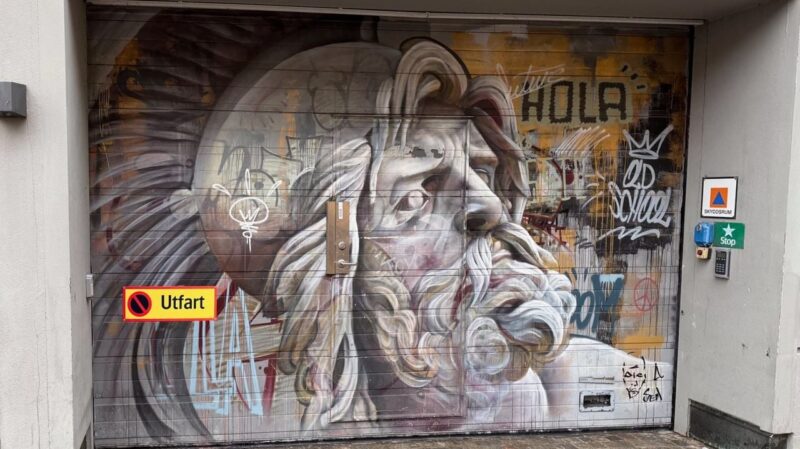 A garage door features a striking mural depicting a side profile of a classical statue with flowing hair and a beard. The artwork is set against a backdrop of graffiti, including tags and the word HOLA. There is a yellow Utfart sign with a red circle and slash, indicating no exit. An access keypad and a blue Stop