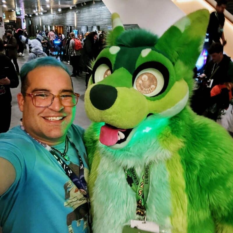 A person with light blue hair and glasses smiles while taking a selfie with a person in a vibrant green and yellow fursuit. The fursuit features large, expressive eyes and a pink tongue sticking out. Both individuals are wearing lanyards with badges. The background shows a busy indoor setting with several people and lights, suggesting a convention or gathering