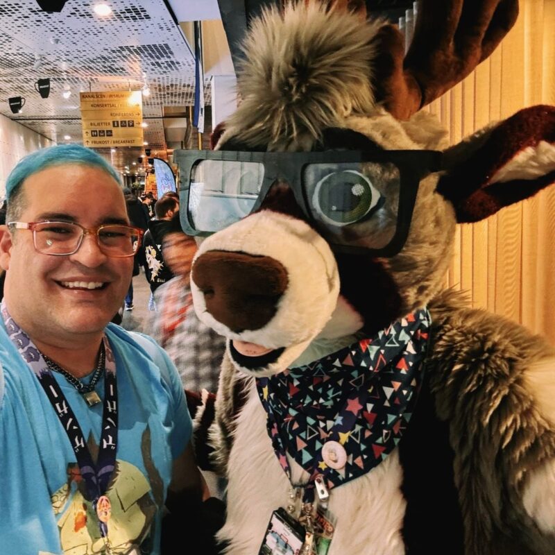 A person with blue hair and glasses is smiling and standing next to a character in a furry costume. The costume resembles a reindeer with large glasses, a fluffy mane, and a colorful bandana with geometric patterns. Both are indoors, likely at an event or convention, as suggested by the signage and other people in the background. The atmosphere appears lively