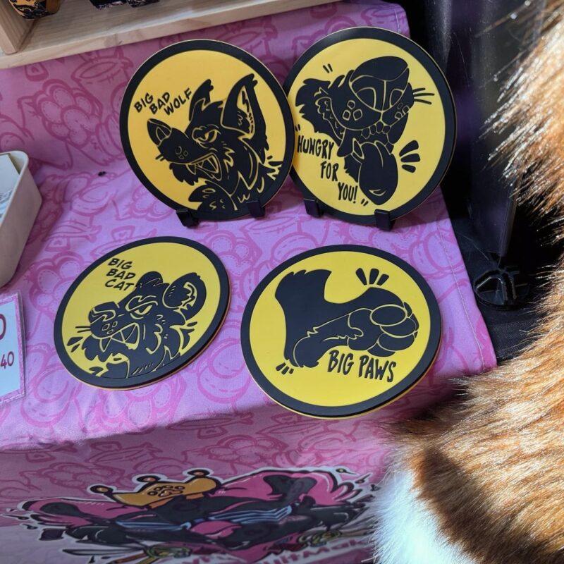 Round, yellow coasters with black cartoon animals and text. Top-left shows a wolf labeled Big Bad Wolf. Top-right features a creature with Hungry for You! Below left is a cat labeled Big Bad Cat. The bottom-right displays a paw with Big Paws. They are on a pink fabric with abstract patterns.