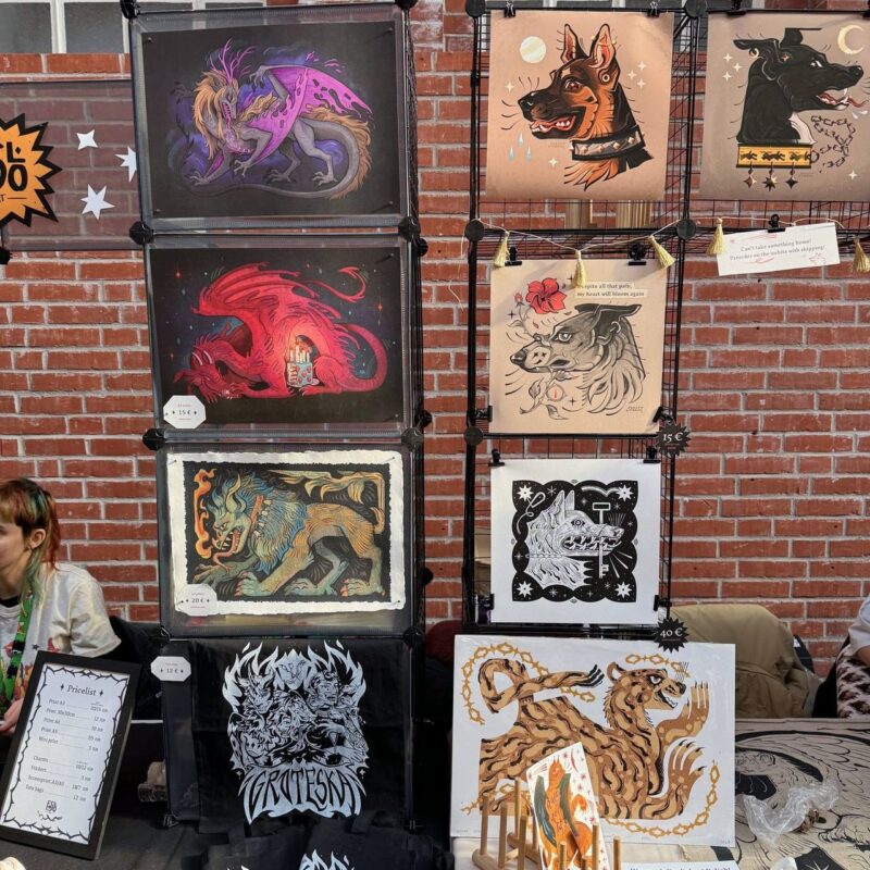 A display of fantasy artwork featuring various mythical and stylized animal illustrations, including dragons, dogs, and wolves. The artwork is mounted on a grid against a brick wall. The images show vibrant colors and intricate designs, such as a dragon with purple wings and a stylized dog with a chain collar. Price tags are visible on some pieces. In the