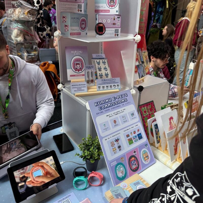 A bustling market stall offering Kurinu e-Pets commissions with merchandise displays. A sign reads “Bring Your Fursona to Life” with customization options for up to four characters. The table holds colorful digital pet devices and wristbands in black, teal, and pink. An iPad shows a close-up of a handheld digital pet. Behind the table,