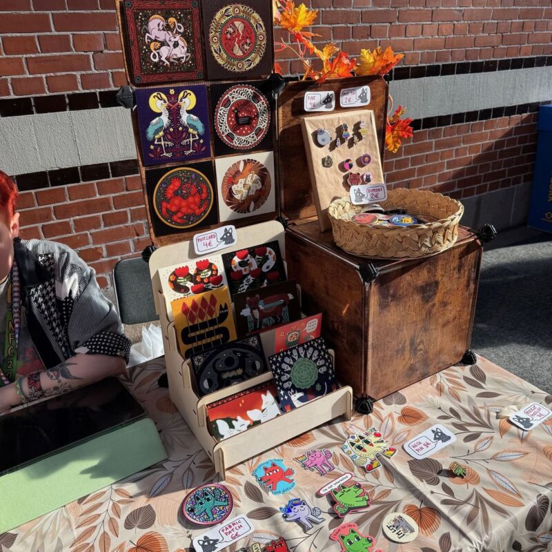 A craft fair booth displays a variety of colorful items on a floral-patterned tablecloth. There are vibrant postcards, pins, and patches featuring whimsical animal designs. Cards are neatly arranged in a small stand, with signs labeled for pricing. A woven basket holds more items. The setup has decorative autumn leaves and a brick wall backdrop. A person with red