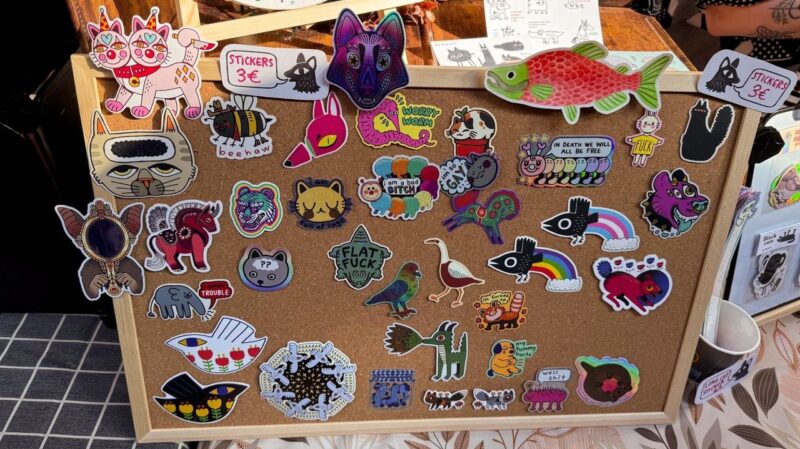 A corkboard displays various colorful and whimsical stickers. The designs include animals like cats, dogs, birds, and fish, as well as unique creatures and abstract shapes. Some stickers have humorous text, such as FLAT FUCK and I am a bad BITCH. The board is labeled with a price: Stickers 3€.