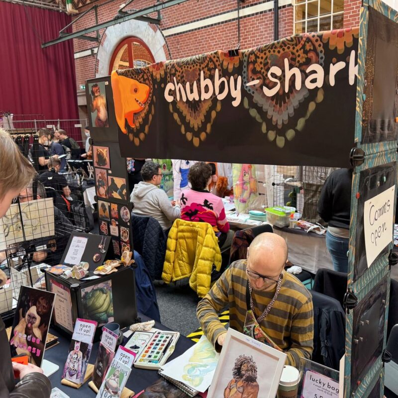The image shows a lively indoor art fair booth. A banner overhead reads chubby-shark, and colorful art prints are displayed on the table and walls. A bald person with glasses and a striped shirt works on an illustration at the table, surrounded by art materials. Various prints and items, such as stickers and keychains, are on display.