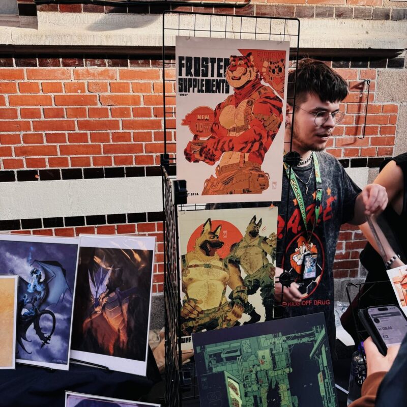A young man stands at a booth with a display of various colorful art prints at an indoor convention. Prominently featured is an illustrated poster of an anthropomorphic tiger in a futuristic outfit, titled FROSTED SUPPLEMENTS. Below it are images of anthropomorphic dogs and mechanical-themed art. The man is wearing glasses and a black shirt