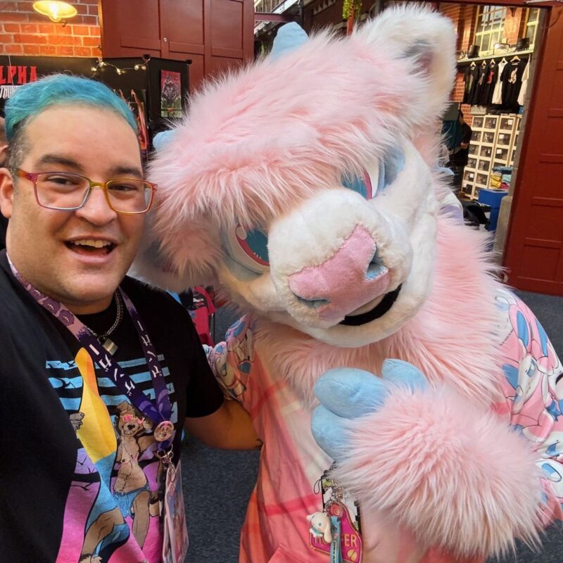 A person with blue hair and glasses smiles while standing next to someone in a pink and white furry costume with a large, plush head and blue eyes. The furry character has a pink and white outfit with a design of cartoon characters and a name badge hanging from their neck. The background shows brick walls and clothing racks in an indoor setting, possibly a convention