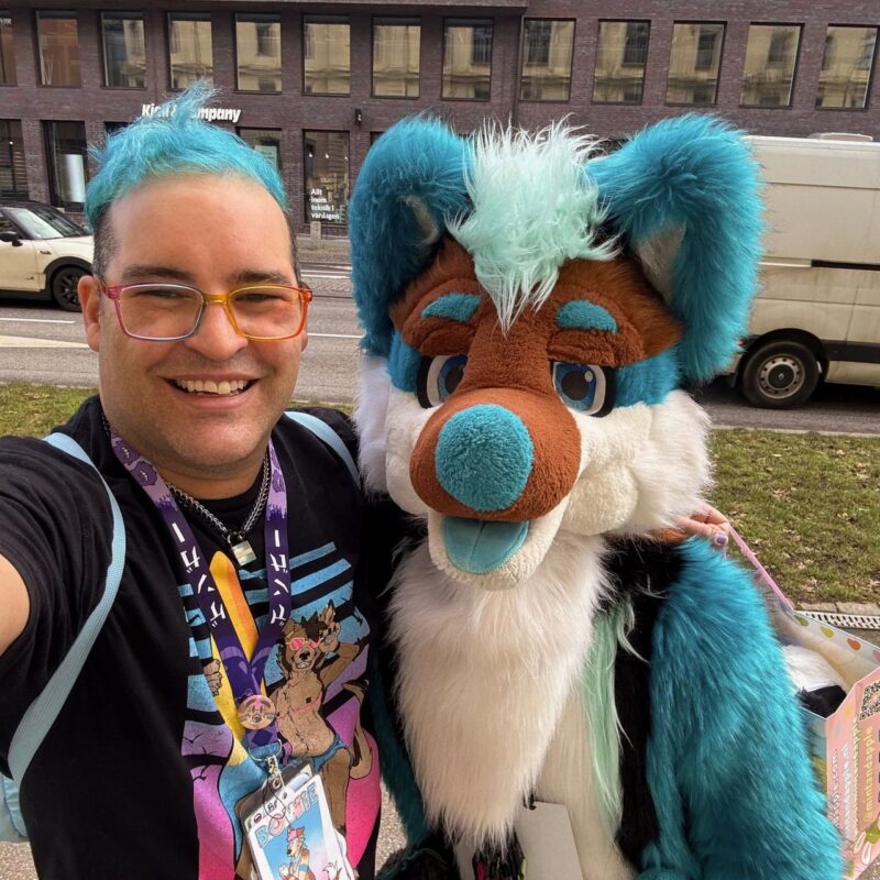A person with blue hair and colorful glasses smiles for a selfie with a person in a furry costume. The costume is vibrant blue and white with brown accents and features large ears and a fluffy mane. The background shows a city street with a brick building and parked vehicles. Both individuals are standing on a grassy area, and the person wears a T-shirt with