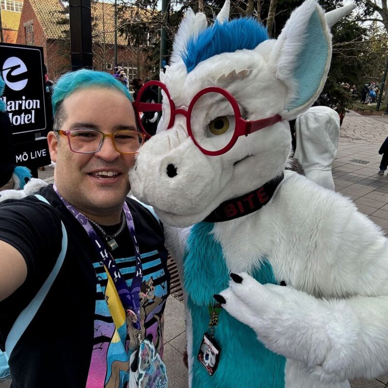 A person with short, turquoise hair and wearing glasses is smiling while taking a selfie. They are dressed in a black shirt with colorful designs and a convention badge on a lanyard. Next to them is someone in a white furry costume with blue accents, wearing red glasses and a collar that reads BITE. The costumed character has large ears