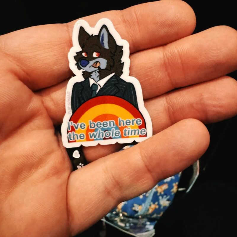 A hand holds a sticker featuring an anthropomorphic wolf character dressed in a suit. The wolf has red eyes, a playful expression, and stands above a rainbow. Below, text reads: I've been here the whole time. The background is blurred, focusing on the hand and sticker.