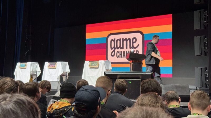 A crowded audience faces a stage set for a Game Changer event. The background displays a vibrant, multi-colored striped pattern with the words game changer in the center. Three podiums covered with white cloth are labeled Erisl, Cashier, and Riff. A man in a black jacket stands to the right