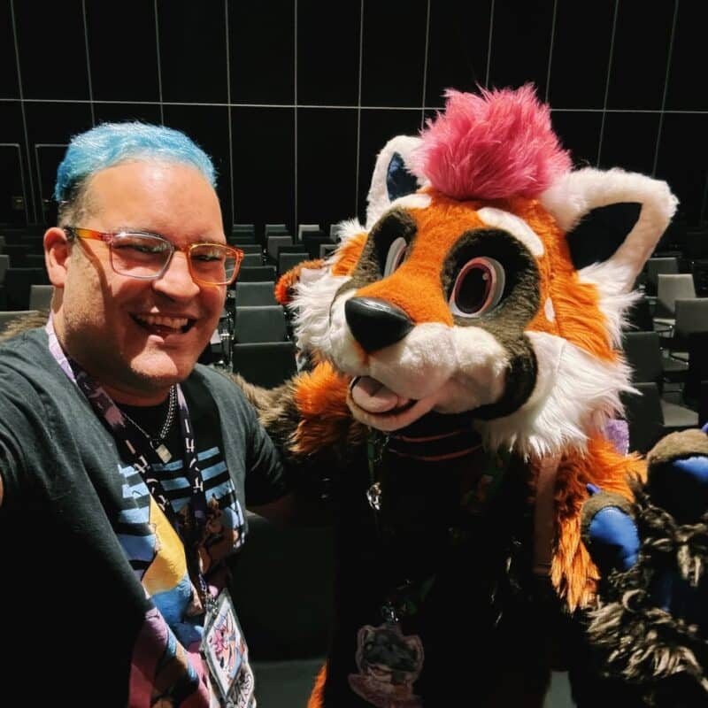 A person with blue hair, wearing glasses and a graphic t-shirt, takes a selfie with someone in a colorful fox-like fursuit. The fursuit features large eyes, a bright pink tuft of hair, and a mix of orange, white, and black fur. They are indoors, with rows of empty black chairs in the background, likely
