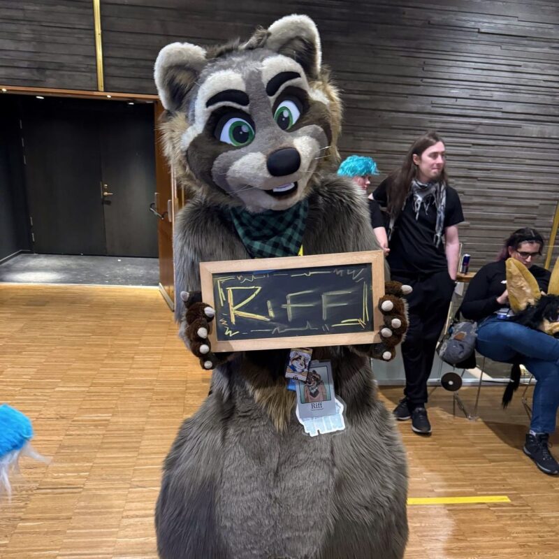A person in a detailed raccoon fursuit, with expressive green eyes and a black-and-green checkered scarf, holds a small sign with the name Riff written on it. The character has a badge with a similar image pinned on. In the background, people are gathered indoors, some sitting and holding a large dog costume head.