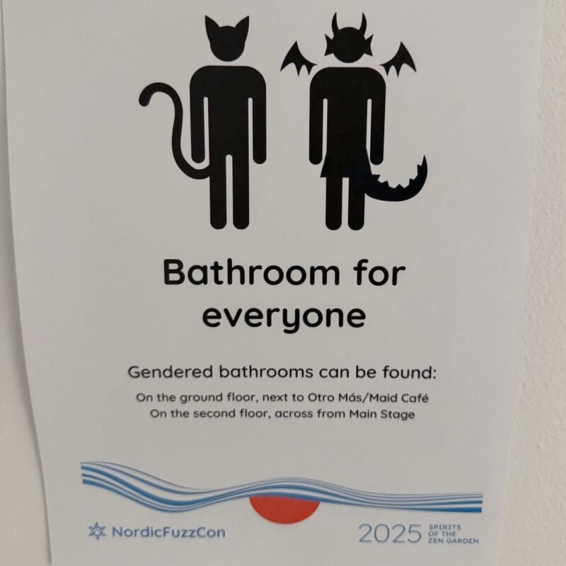 A sign depicting two stylized figures, one with cat ears and tail and the other with horns, wings, and a tail. The text reads Bathroom for everyone with additional information stating that gendered bathrooms are available on the ground floor next to Otro Más/Maid Café and on the second floor across from Main Stage. At the bottom, it