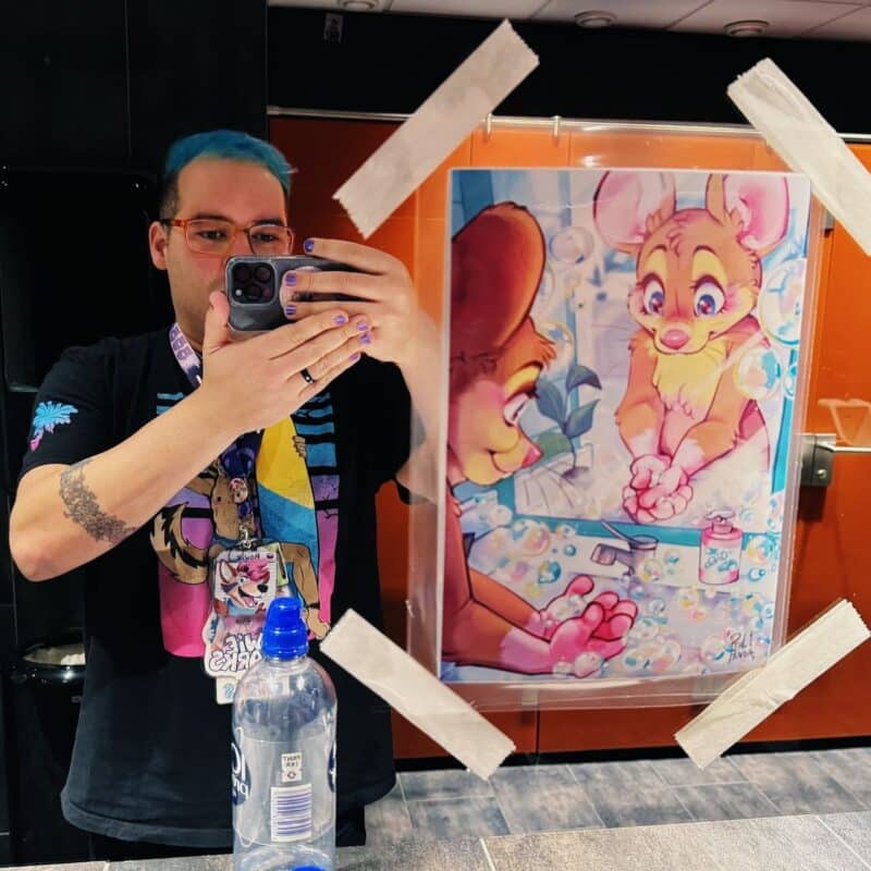 A person with blue hair and glasses takes a selfie in a bathroom mirror. They are wearing a black T-shirt featuring a colorful cartoon design and a lanyard with a badge. Their nails are painted purple. In front of the mirror is a taped-up poster showing a cartoon-style illustration of a cute animal washing its hands with soap bubbles. A water