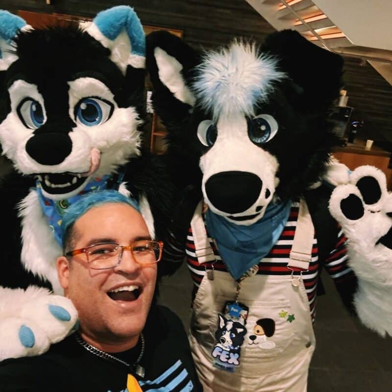 A person with blue hair and glasses is smiling broadly while taking a selfie with two individuals in black and white furry costumes resembling dogs or wolves. The costumes feature large blue eyes and distinct blue accents. One costume has a blue handkerchief with colorful patterns, while the other wears a striped shirt and overalls with a blue bandana. Both costume wear