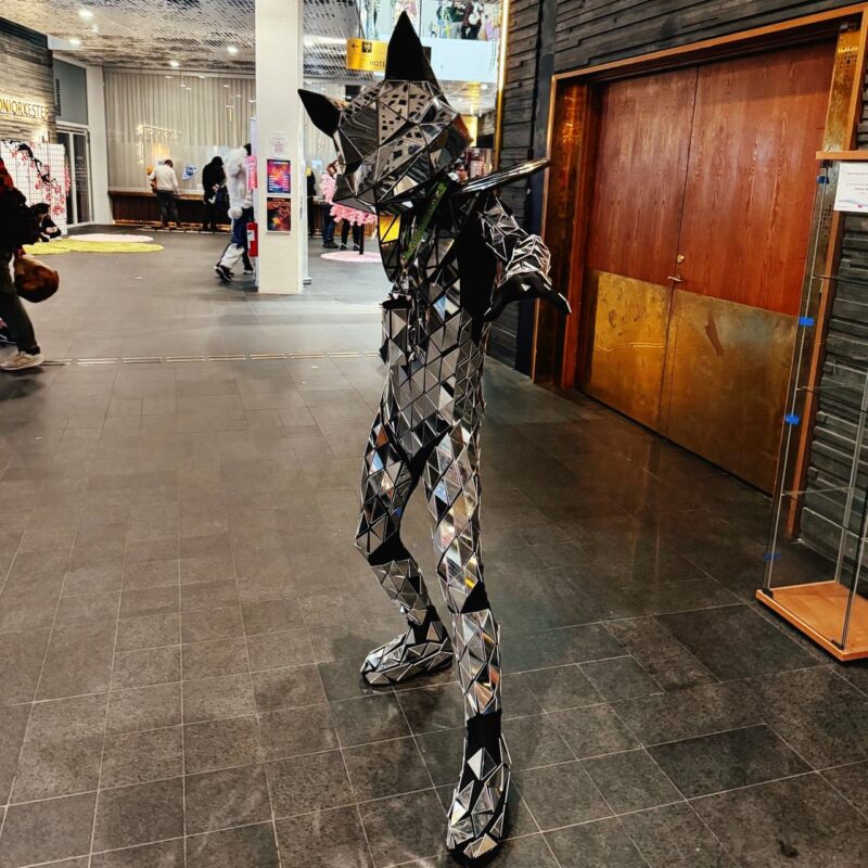 A person is dressed in an elaborate costume resembling a cat, composed of reflective, geometric, mirror-like panels. The outfit is predominantly silver, creating a faceted texture that catches the light. The setting appears to be indoors, possibly a convention or event space, with people and displays visible in the background. The floor is tiled, and the decor is