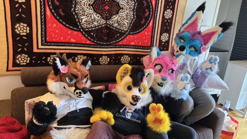 Four individuals are wearing colorful fursuits, sitting on a sofa. The costumes include a brown fox, a black and yellow dog, a pink and blue wolf, and a blue, white, and purple fox. Each character has distinct, expressive eyes and matching paws. They sit in front of a detailed tapestry with floral and geometric patterns in red,