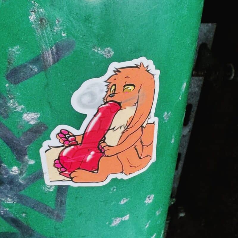 A sticker on a green surface depicts a cartoonish, orange-furred creature sitting with a relaxed expression. The creature is holding a large, red, heart-shaped object. Its ears are long and floppy, and its eyes are half-closed. The sticker stands out against the textured, slightly worn-looking green background.