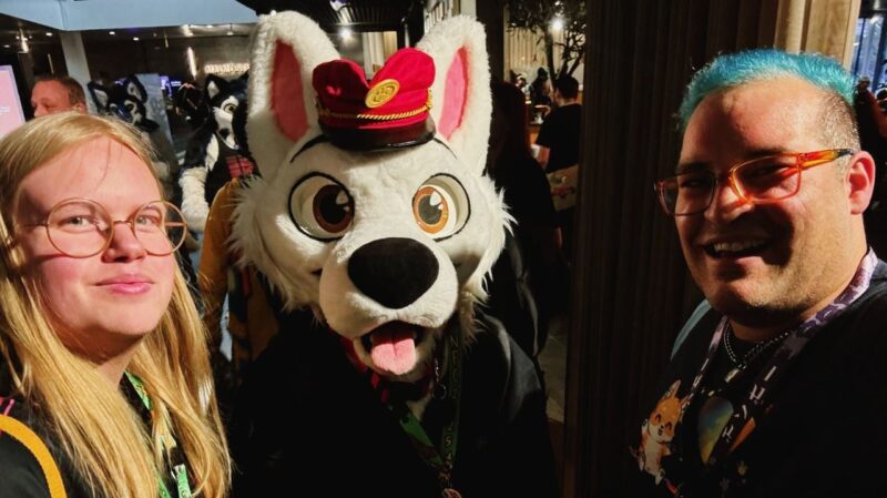 Three people are posing for a photo at a lively indoor event. In the center, a person is wearing a white dog costume with a red hat, featuring large expressive eyes and a sticking out tongue. They are flanked by two smiling individuals wearing glasses; one has long blonde hair, and the other has short, bright blue hair. Both have l