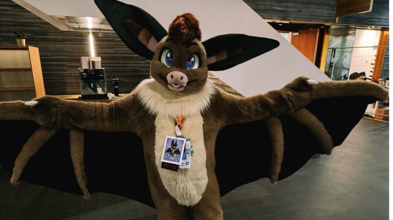 A person is wearing a brown bat fursuit with large ears and blue eyes. The suit has outstretched wings and a light-colored furry chest. They are in an indoor setting with modern decor, including shelves and an espresso machine in the background. The person is wearing convention badges on a lanyard around their neck. The scene gives a