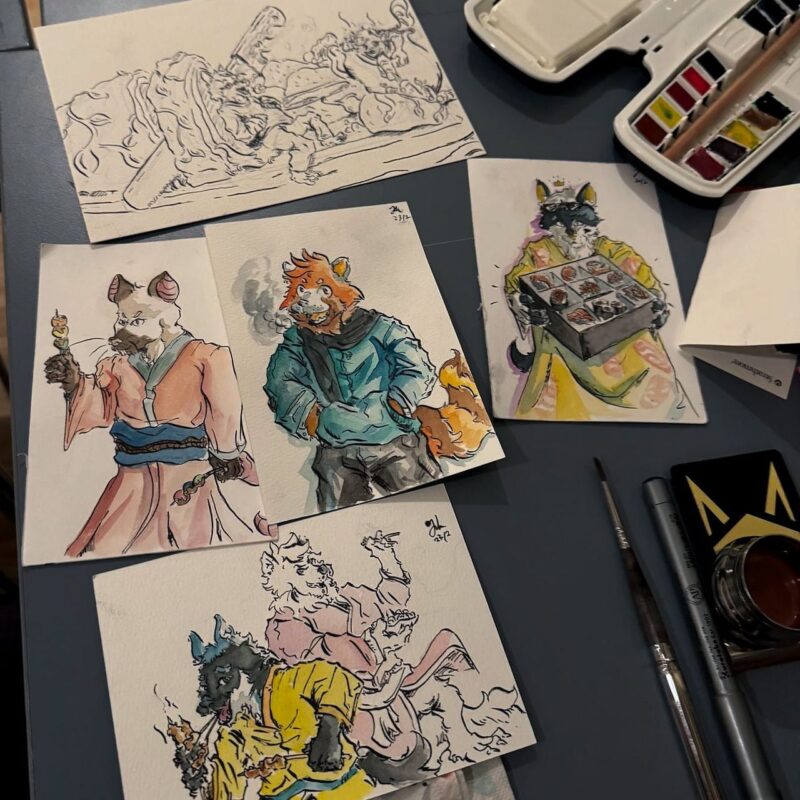 This image displays a collection of watercolor paintings and drawings featuring anthropomorphic animal characters. One character resembles a fox in a light blue jacket, another is a raccoon carrying a sushi tray, and a third is a wolf in robe-like attire. The artworks are laid out on a table alongside painting supplies, including a watercolor palette, brushes, and a small