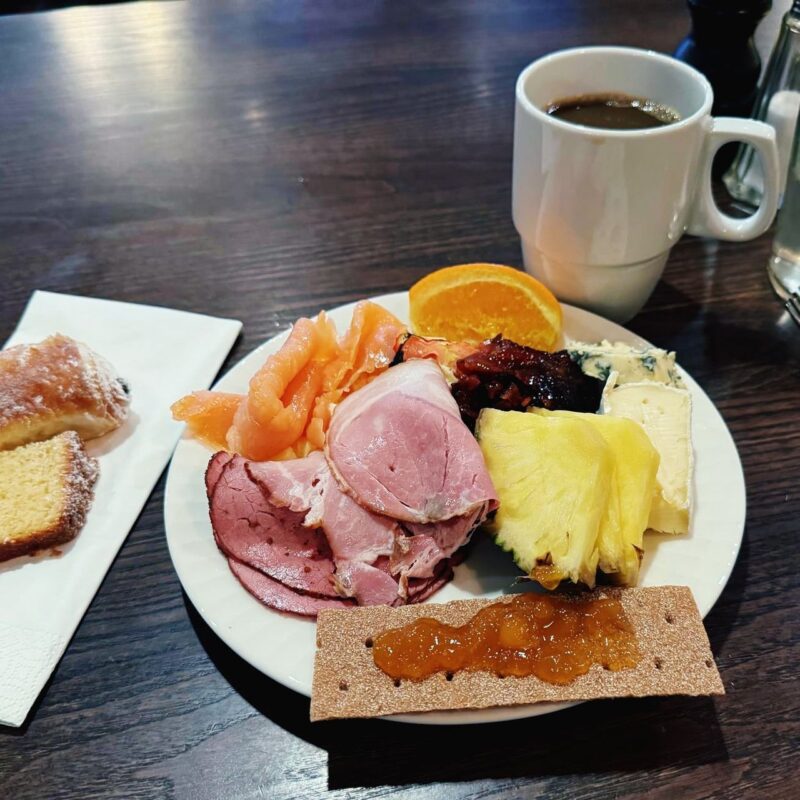 A plate filled with a variety of breakfast items including slices of ham, smoked salmon, orange, roasted meats, blue cheese, brie, pineapple, and a cracker topped with jam. Next to the plate is a cup of coffee. To the side, there’s a croissant and a piece of pound cake on a napkin. The setting is