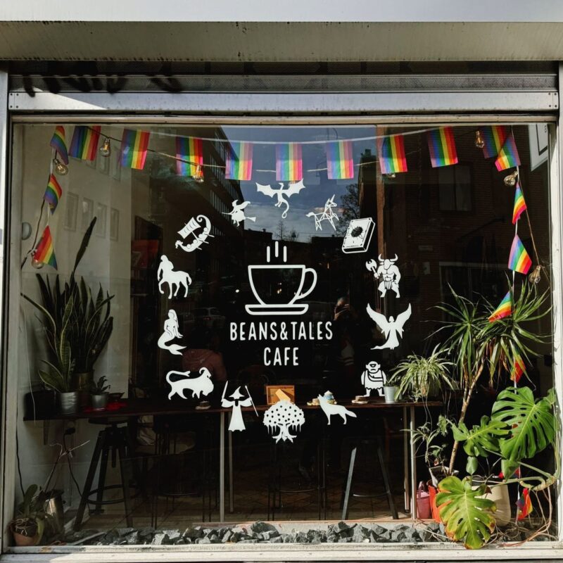The image shows the storefront of Beans & Tales Cafe. The window features white decorative illustrations of mythical creatures and symbols surrounding a steaming coffee cup logo. Colorful rainbow flags drape along the upper edge of the window. Inside, visible are tables with patrons, a cozy interior, and potted plants. The scene reflects a welcoming and creative atmosphere, blending