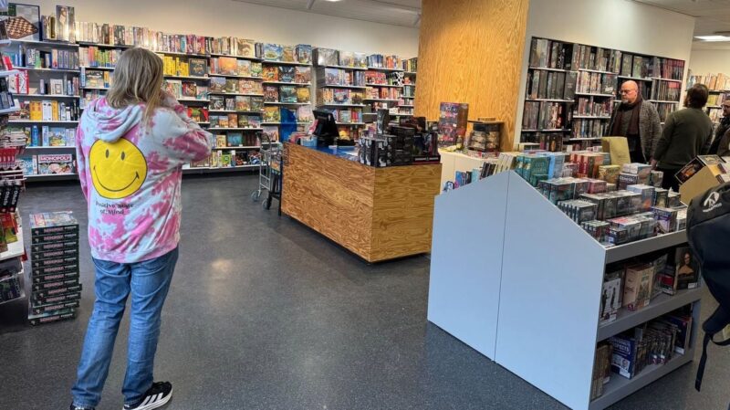 A person wearing a pink and white tie-dye hoodie with a large yellow smiley face and the text Positive State of Mind stands in a board game store. They face a wooden checkout counter with various game boxes on display. Shelves filled with board games, including Monopoly, line the walls. Two people are visible in the background, browsing