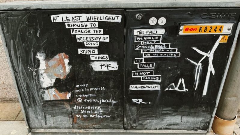 A graffiti-covered metal box featuring various white text snippets on a black background. The left side reads, At least intelligent enough to realise the necessity of doing stupid things, with an illegible signature below. The right side displays phrases and words like The fall, It falls in not daring, and Vulnerability, alongside an illustration of a