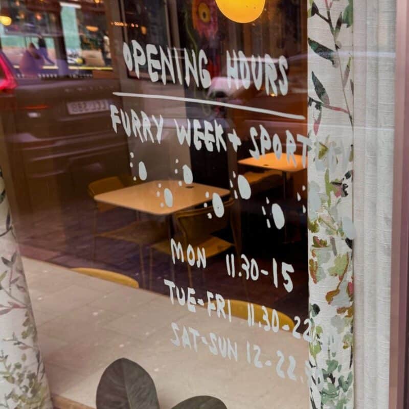 The image shows a glass door with written text displaying the opening hours of an establishment. It reads: Opening Hours Furry Week + Sport, followed by paw print designs. The detailed hours are: Monday 11:30-15:00, Tuesday-Friday 11:30-22:00, and Saturday-Sunday 12:00-