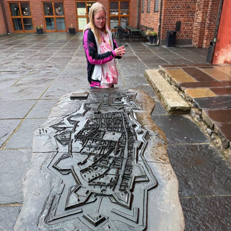 A person wearing glasses, a pink and white tie-dye shirt, and a black and pink jacket is standing in a courtyard. They are observing a detailed topographical bronze model of a historic town or fortress placed on a stone table. The smooth, wet surface of the model suggests it has recently rained. Behind the person, there are brick buildings