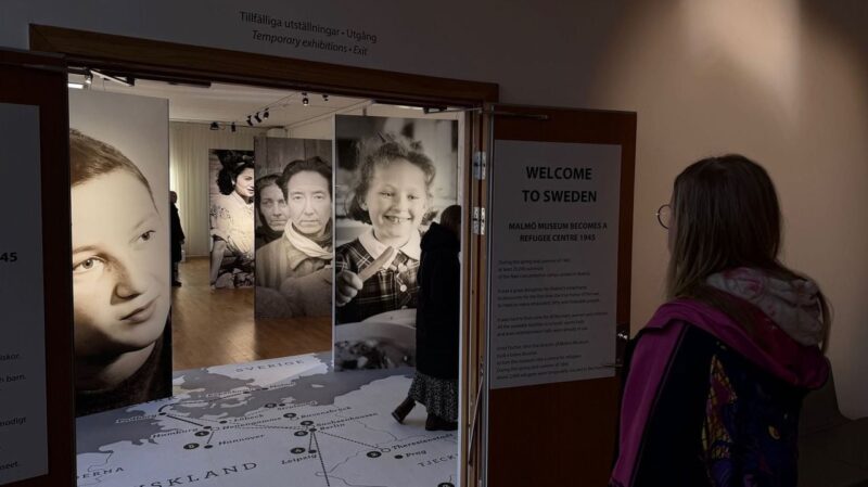 A person in a colorful jacket and glasses stands in front of a doorway at an exhibition, titled WELCOME TO SWEDEN. The text explains Malmö Museum's role as a refugee center in 1945. Inside, large black-and-white photos of diverse people, including children and adults, are displayed. The floor features a detailed map, likely depicting
