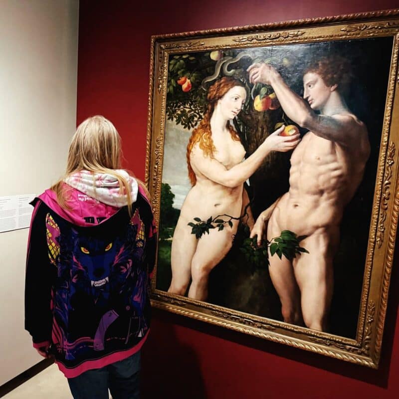 A person with long blonde hair, wearing a black jacket with a colorful wolf design, stands in front of a large framed painting hanging on a red wall. The painting depicts a scene from the biblical story of Adam and Eve, featuring two nude figures with an apple tree and a serpent. The person is viewing the artwork attentively, creating a contrast between