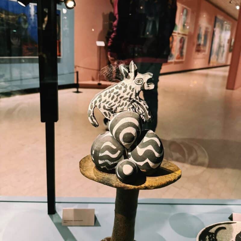 The image shows a ceramic sculpture displayed in a museum setting. The sculpture features a small, abstract animal figure standing atop a stack of eggs with wavy, zebra-like patterns. The pedestal supporting the figure is round and textured. The background includes blurred artwork on peach-colored walls, with a person faintly visible reflecting in the glass display case. The lighting