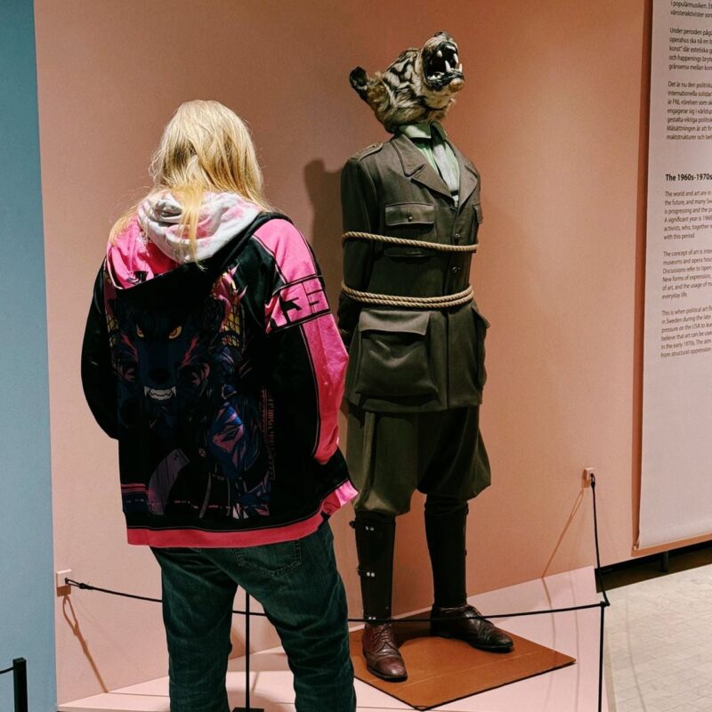 A person with long blonde hair, wearing a colorful hoodie featuring a wolf design and jeans, stands in front of an art installation. The installation includes a mannequin in a military uniform with a roaring animal head and a rope tied around its torso. The background consists of light pink walls with text panels. The scene appears to be in a museum or gallery setting