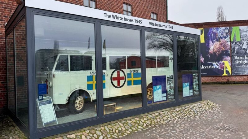 A white historic bus displayed in a glass enclosure outside a brick building. The bus features Swedish flags and a red cross on its side, with the text Sverige and Schwi visible. A sign at the top reads The White buses 1945 and Vita bussarna 1945. Informational panels are placed