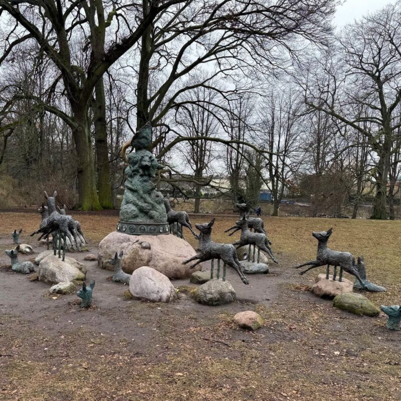 A dynamic statue garden features sculptures of wolves mounted on short poles, giving the appearance they are leaping over scattered rocks. In the center, there is a larger sculpture of a mystical figure or creature, surrounded by the wolf sculptures. The ground is bare, with a few patches of grass, and the background is filled with leafless trees, suggesting a
