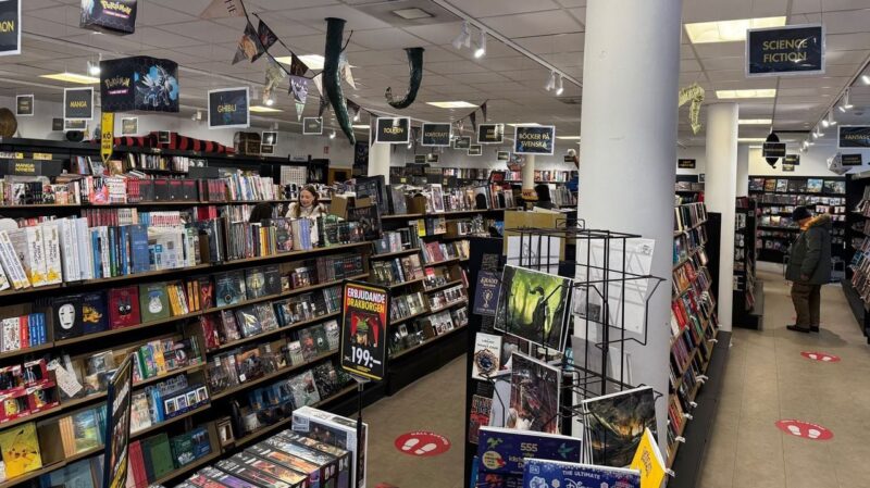A bustling bookstore interior with several aisles lined with shelves of books and comic books. Overhead, signs categorize sections like Science Fiction, Manga, and Ghibli. The aisles also feature suspended decorations, including tentacle shapes and paper flags. A customer in a green coat browses near the back. The floor has social
