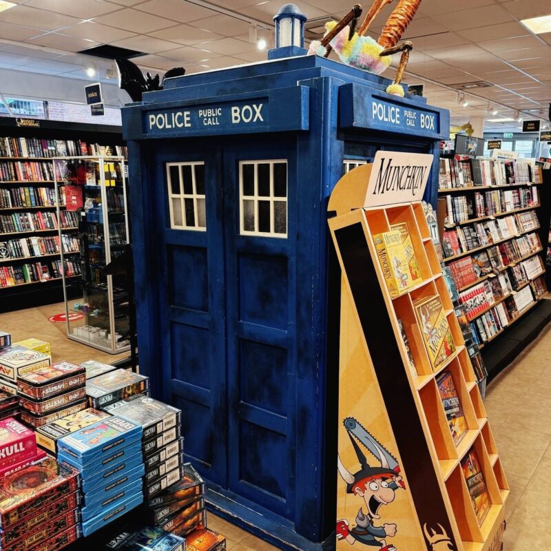 A life-sized replica of a blue police box, resembling the TARDIS from Doctor Who, stands in a bookstore amidst shelves lined with books. In front of the TARDIS, there is a display for the game Munchkin, with various editions stacked neatly. The store’s interior features bright lighting and well-organized shelves filled with
