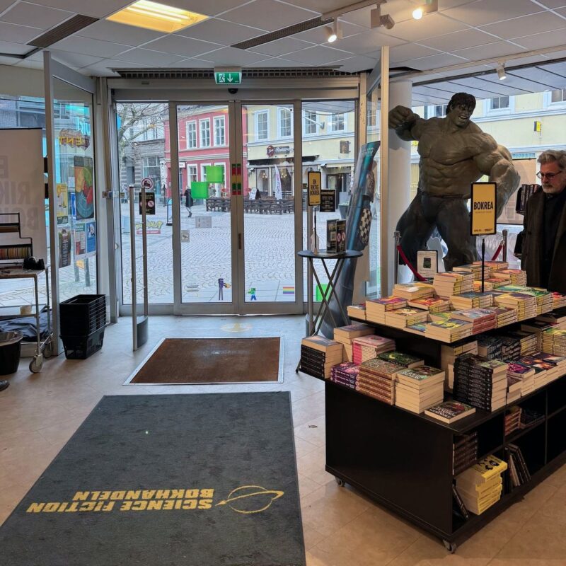 A bookstore entrance with large glass doors provides a view of a cobblestone street and colorful buildings outside. Inside, a life-sized Hulk statue stands prominently near book displays filled with various titles. A tall man wearing a black coat browses the selection. The floor has two mats, one with the words Science Fiction Bokhandeln printed on it