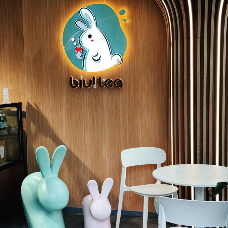 A modern tea shop interior with a wooden wall featuring a glowing logo of a rabbit holding a drink and the text biu!tea. There are two rabbit-shaped chairs, one mint green and the other pink, along with a round white table and chair. Vertical wooden slats with lighting add a stylish touch to the right side of the image,