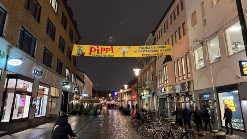 A cobblestone street at night, lined with shops and softly lit by street lamps. A yellow banner hangs overhead, promoting Pippi Långstrump with images of the character and a horse, announcing a premiere on March 22 at Nöjesteatern, Malmö. People walk along the street, some with bicycles parked nearby.