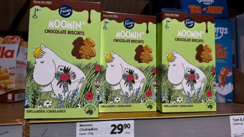 Three boxes of Moomin chocolate biscuits are displayed on a shelf. The packaging features a cartoon Moomin character holding a colorful bouquet of flowers against a green background with grass and flowers. A small image of a chocolate biscuit shaped like a character is shown on the box. The product is marked as palm oil-free and states Responsible Cocoa 100