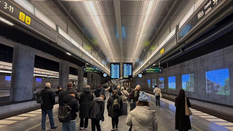 A bustling train station platform with people standing and walking, many wearing winter clothing. The ceiling is lined with bright fluorescent lights and metallic panels. Above the platform are digital displays showing train schedules. The walls feature large digital screens displaying various images, possibly advertisements or art. The platform design is modern and spacious with concrete pillars and tiled flooring.