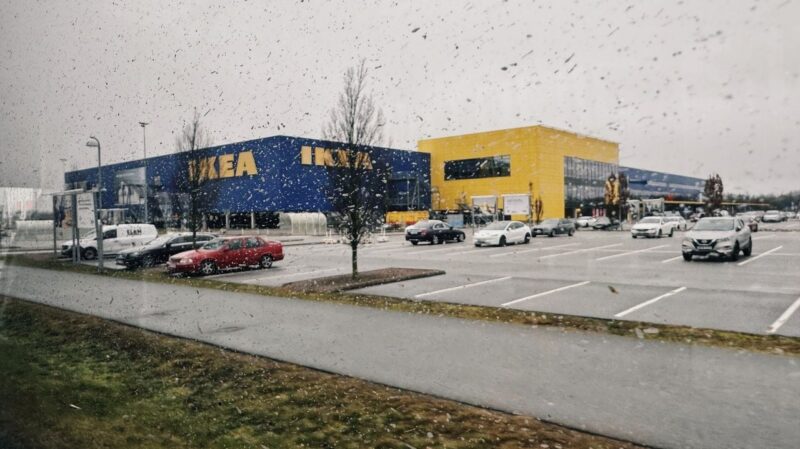 The image shows a large IKEA store with blue and yellow exterior walls, seen through a rain-speckled window. In front of the store, there is a spacious parking lot with several parked cars, including a red sedan and a white SUV. The sky is overcast, contributing to a gray, dreary atmosphere. Leafless trees are scattered around