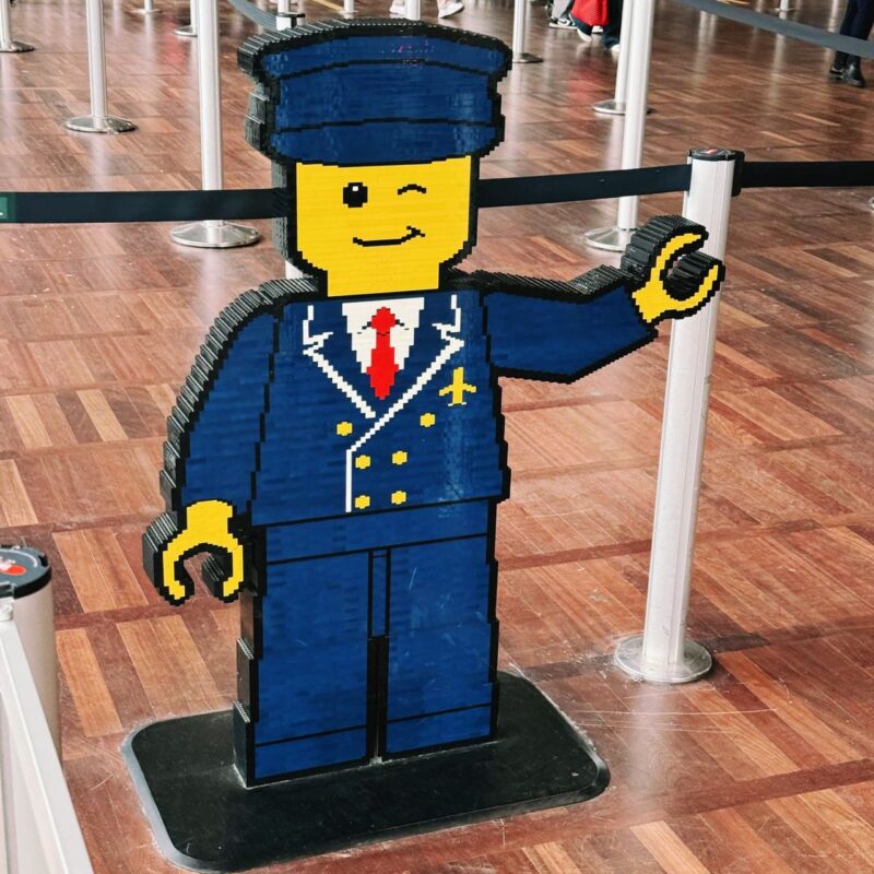 A life-sized LEGO-style sculpture of a pilot stands on a wooden floor. The figure is dressed in a blue uniform with a red tie and gold details, including buttons and an emblem. It is winking and raising its left hand in a greeting gesture. The sculpture is surrounded by black retractable barriers, typical of queue lines in public places, like