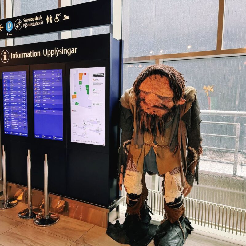 A large puppet resembling a troll is positioned to the right of an information board inside a building. The board displays travel or service information in both Icelandic and English, with digital screens showcasing schedules and maps. The troll figure is dressed in mismatched, tattered clothing and has a rugged, bearded appearance. The setting seems to be an airport or